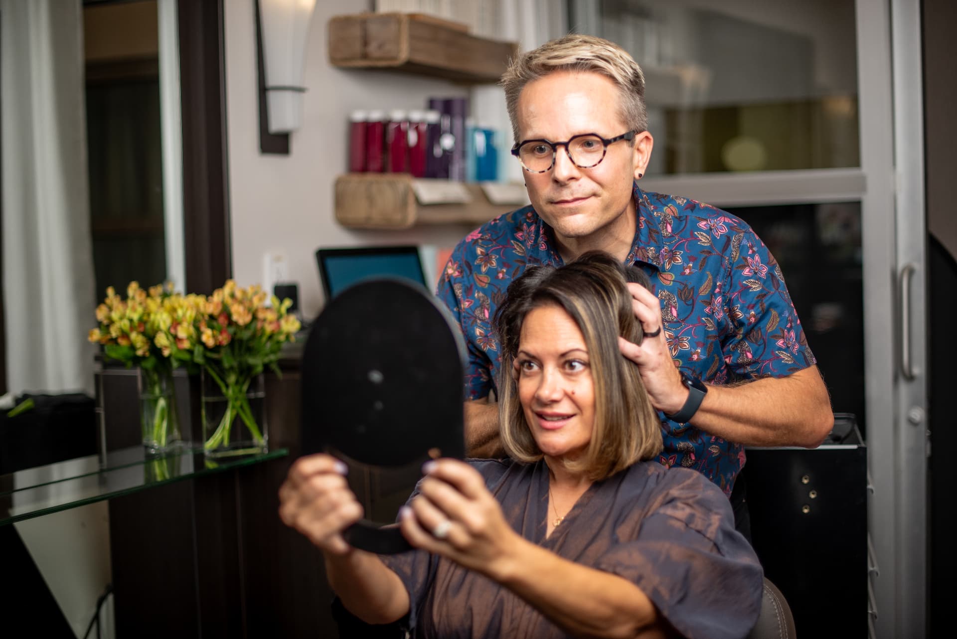 A delighted client checking her fresh color in a hand mirror while Ray lifts a section of her hair.